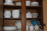 Full view of cupboard shelves displaying stacked Corelle white plates and bowls, colorful Cordella stoneware plates, and white ceramic tea pots and napkin holder on lower shelf.