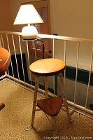 Wood and metal stool with round wooden seat, triangular flip-down step, and distressed white legs shown next to a white ceramic touch table lamp with a cream-colored square shade and visible damage.