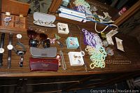 Photo shows a variety of costume jewelry including beaded necklaces, brooches, watches, sunglasses, and small boxes on a wooden dresser surface