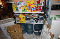 Photo showing assorted laundry and cleaning supplies on shelf including Tide detergent, Glad bags, Clorox bleach, Black and Decker iron, SOS pads, Resolve spray, multiple other bottles, and several blue water jugs stored beneath shelf along with laundry hampers.