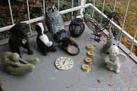 Photo showing twelve outdoor garden decor items arranged on porch floor, including animal figures, an angel statue, a thermometer, and four small weather instruments with wear.