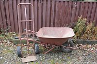 Metal dolly beside metal wheelbarrow outdoors on gravel, showing both items from side angle, highlighting wear and condition.
