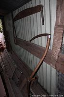 Long two-person saw hung horizontally on a metal wall with a curved wooden scythe positioned underneath, wooden planks stacked below.