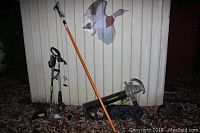 Black and Decker hedge trimmer, Earthwise trimmer/edger, LawnMaster leaf blower and orange tree pruner pole leaning against a metal shed outdoors on leaf-covered ground.
