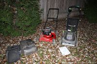 Two lawn mowers side by side outside on leaf-covered ground near a wood fence. Grass collection bags and some documentation are shown on the ground.