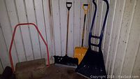 Three snow shovels and a roller seen against a corrugated wall inside a shed, showing the condition and arrangement of all items.