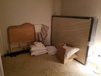 Full view showing queen size mattress, boxspring, black metal bed frame, wicker headboard, and bedding piled on floor.