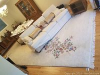 Wide view of large white wool area rug with fringed edges and floral center design in a living room setting beneath a sofa and dining table.