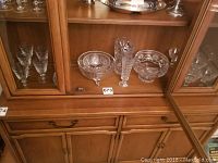 Wide view of crystal and glass items inside wooden cabinet including serving bowls, footed condiment dishes, glass stir sticks, and wine and dessert glasses.