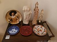 Photo of a wooden table with three decorative plates and three statues including a classical statue torso, Asian warrior statue, and a standing Buddha statue.