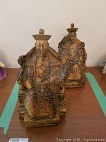 Photo showing two Asian ceramic figurines of a King and Queen with detailed robes and headpieces seated on chairs.