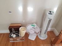 Wide view of the lot showing Nordic tower fan, Westinghouse table fan, bath mat, caddy with hairdryer and towel on hardwood floor against white wall.