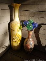 Tall yellow ceramic vase with floral motif and handles, standing next to a smaller patterned vase with glass water globes.