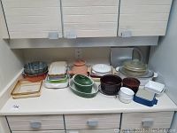 View of countertop with assortment of bakeware including two Le Creuset casserole dishes (brown and green), Pyrex glass baking dishes, ceramic baking dishes, ramekins, and metal bundt pan.