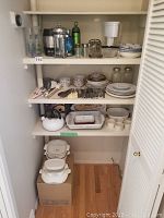 Wide view of kitchen pantry shelves containing assorted kitchenware: plates, bowls, glass jars, cutlery, CorningWare dishes, white serving dish, and white kettle.