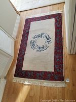 Photo of the entire rug laid out on a hardwood floor, showing the cream center with a blue circular design and red patterned border with fringes.