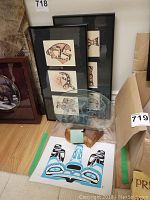 Two framed black and red Native art prints leaning against wall with blue and black print and etched glass nearby
