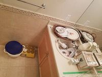 Corner floor area and bathroom sink counter showing stacked Royal Doulton plates, an oval serving platter, small metallic bowls and bowls in a basket, next to an electric hand mixer and white pitcher.