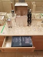 Overview of multiple crystal and glass drinkware including wine glasses, snifters and boxed items on countertop and drawer.