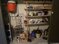 Photo showing shelves with assorted garden pots and various garden tools placed beneath them along with wire plant supports and baskets.