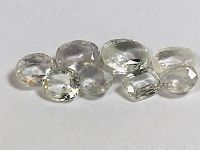 Eight faceted white sapphires with varying shapes and sizes, clear to slightly off-white color, visible natural inclusions, against white background.