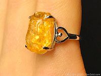 Close-up detail of the yellow citrine carved stone set in a silver ring worn on a finger, showing the translucent warm yellow color and the carving details.