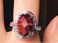 Close-up of the ring showing the faceted oval red tigers eye gemstone and the decorative silver bezel with Greek key pattern. Ring is worn on finger, showing adjustable band.