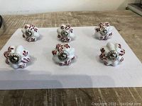 Top view of six ceramic knobs arranged in two rows on a white sheet, showing the red leaf designs and silver metal screw centers.