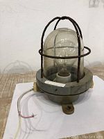Rustic antique ship light with glass dome and protective metal cage on wooden surface, showing wiring and base.