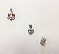 Three silver pendants displayed on a white background: flower pendant with multi-colored stones, heart pendant with green stones, swirl pendant with yellow stone