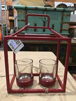 Red metal house-shaped frame with two clear glass holders positioned inside on a surface with vintage trunks in the background.