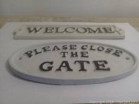 Photo showing two white metal plaques with raised bronze-colored letters viewed from above on a wood surface, one rectangular "WELCOME" and one oval "PLEASE CLOSE THE GATE" sign.