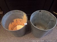 Top down view showing the interior and exterior of two vintage galvanized buckets placed side by side on a carpeted floor, highlighting their condition and size