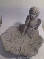 Grey cupid figure sitting on edge of flower-shaped bird bath basin.