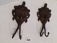 Front view of two double hooks showing detailed handcrafted brass face designs with ornamental features and twin hooks extending downwards.