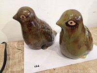 Two ceramic birds with a glossy green and brown glaze, showing rounded body shapes and sculpted facial and wing details, displayed on white paper.