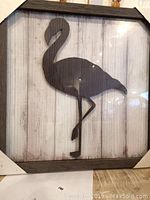 Front view of framed crane silhouette artwork showing bird standing on one leg on a white plank background with dark wooden frame and glass front.