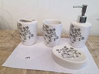Four white ceramic bathroom vanity accessories with black floral design on a wooden surface: soap dispenser with pump, toothbrush holder, rinsing cup, and soap dish.