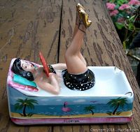 Front-left angle view of the porcelain figural ashtray showing bathing beauty lady with raised legs, black polka dot swimsuit, golden heels, and tropical decoration sides with flamingos and palm trees.