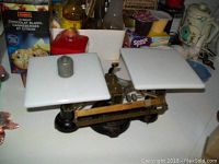 Front angled view of the black japanned metal OHAUS balance scale showing the left porcelain plate and a metal weight on top.