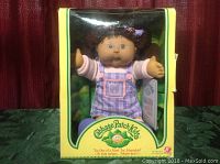 Front view of boxed Juanita Vivica Cabbage Patch Kids doll in yellow Play Along 2004 packaging, showing doll with arms extended, purple plaid jumper with pink shirt, and butterfly patch.