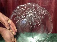 Round clear glass serving platter with embossed Christmas bells and holly decoration held by hands, showing front detail and size reference.