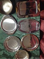 Top view of five silver trays and two silver bowls arranged on green fabric showing variety of shapes and sizes.