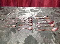 Full view of 15 pieces: 13 collector teaspoons and 2 miniature forks on green fabric background.