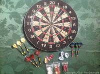 Dartboard with nine darts arranged around it and additional flights in front on a table