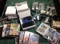 Overview of the gaming lot showing PS Vita handheld console in box, Nintendo Wii console with cables, assorted game discs mostly PlayStation 2 and Wii titles, and multiple controllers laid out on a green surface.