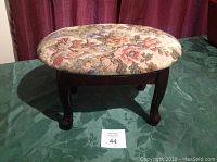 Front view of oval vintage upholstered footstool with floral fabric and wooden cabriole legs on green surface