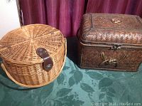 Two wicker baskets side by side on a green cloth, a round natural wicker basket with a leather strap clasp on the left, and a rectangular darker woven basket with a latch on the right.