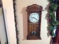 Front view of vintage wooden wall clock with carved top, white clock face, floral glass panel, and visible pendulum inside case.