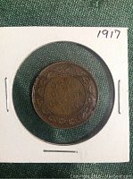Photo showing the reverse side of the 1917 Canadian penny with denomination and date surrounded by a wreath, mounted in a white cardboard coin holder.